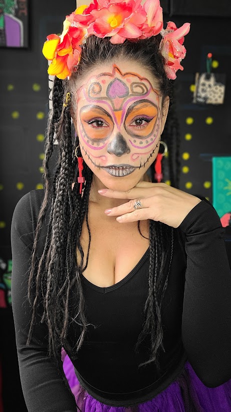 Calavera orange et mauve avec cœur et spirales sur le front, longues tresses et couronne de fleurs chaudes.