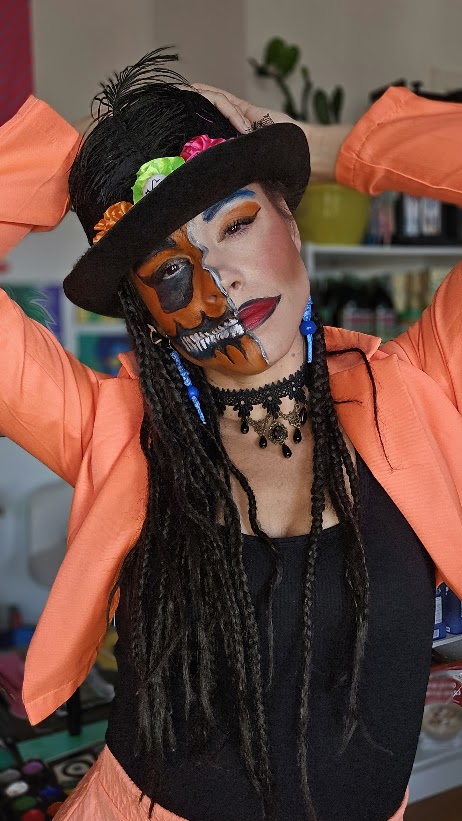 Demi-squelette orange et noir avec dents blanches, maquillage glamour de l’autre côté et grand chapeau noir fleuri.