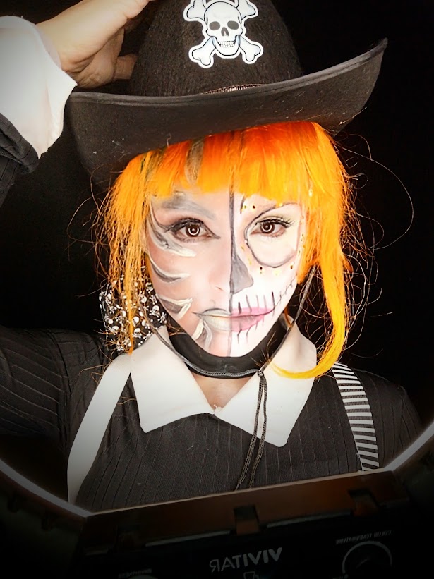 Maquillage asymétrique argenté et blanc avec demi-sourire de squelette, cheveux orange et chapeau noir.