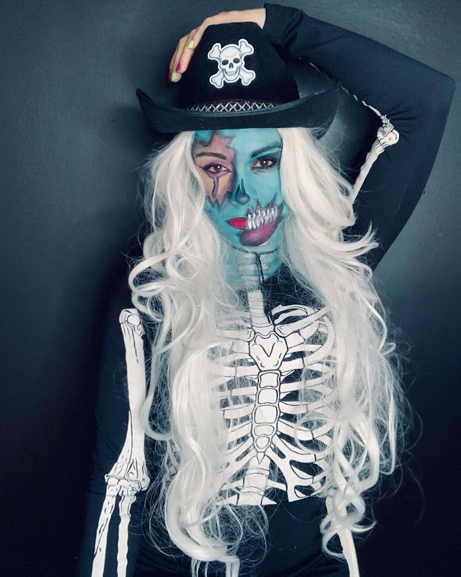 Maquillage turquoise avec zone rose autour d’un œil et dents de squelette, perruque blanche et chapeau noir.